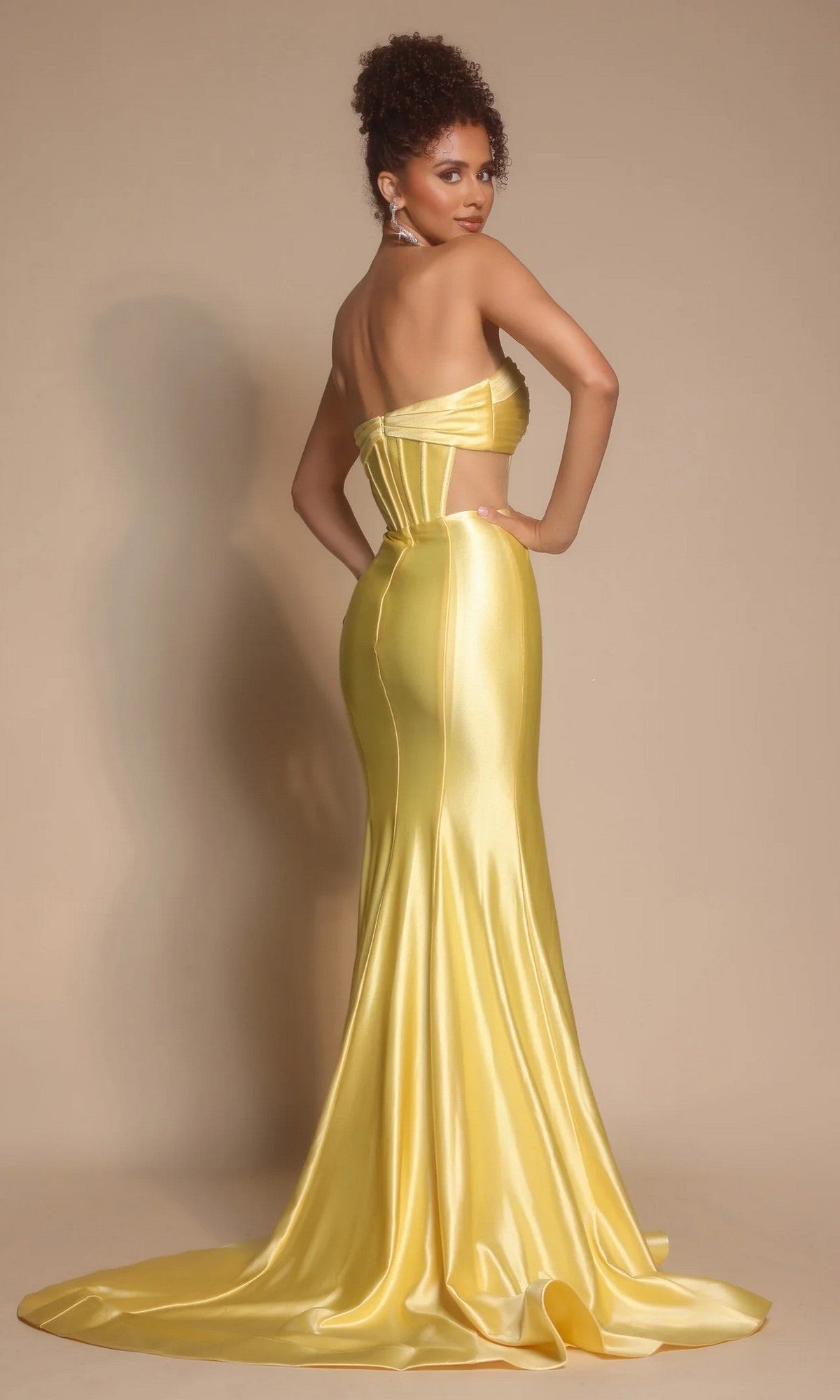 A woman faces backward, looking over her shoulder in the Portia and Scarlett XO066 long formal dress featuring a strapless satin design with a pleated crossover bodice and a flowing train.
